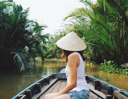 A woman floating down the Mekong River facing away from the camera wearing a traditional nón lá hat