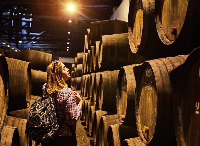 Une jeune femme blonde contemple des tonneaux en bois dans les caves d'un domaine viticole.