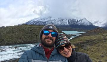Two people smiling outdoors with scenic mountains and a river.