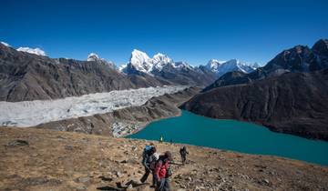 Hikers descending a mountain path with a view of a glacier and lake.