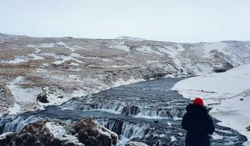 Person in winter attire standing by a snowy river with mountains.
