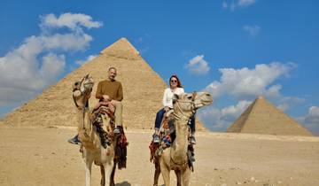 Couple riding camels in front of pyramids.