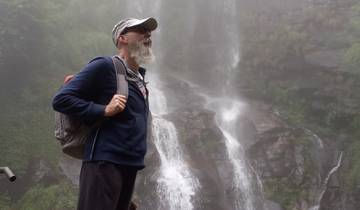 A man with a beard standing near a waterfall in a misty environment.