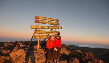 Two people posing at Uhuru Peak with Kilimanjaro summit signs.