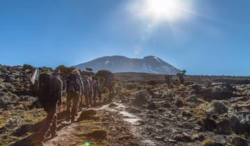 Hikers on a trail with Mount Kilimanjaro in the background under a bright sun.