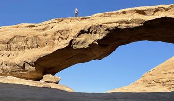 Person standing on top of a large natural rock arch in a desert landscape.