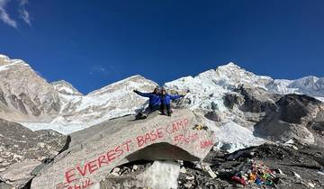 Two people on a rock with Everest Base Camp sign, snowy mountains in the backdrop.
