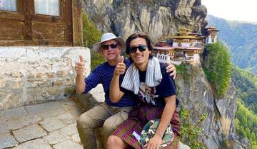 Two people posing with thumbs up near a cliffside monastery.