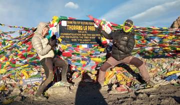 Two people posing with a sign at Thorong La Pass, surrounded by prayer flags.