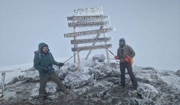 Two people at a snowy mountain summit with signage.