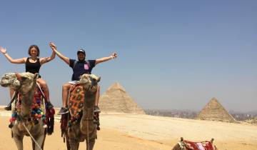 Two people riding camels with the Pyramids of Giza in the background.