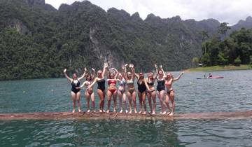 Group of women in swimsuits waving beside a lake.