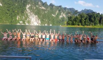 A group of people sitting on a dock with their hands up, with mountains and a lake in the background.