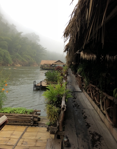 Riverside with mist and traditional huts along a walkway.