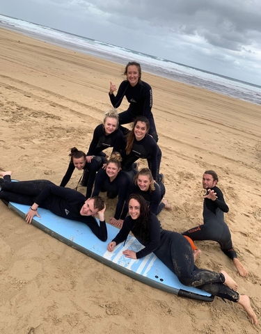 Group of people in wetsuits posing on the beach with a surfboard.