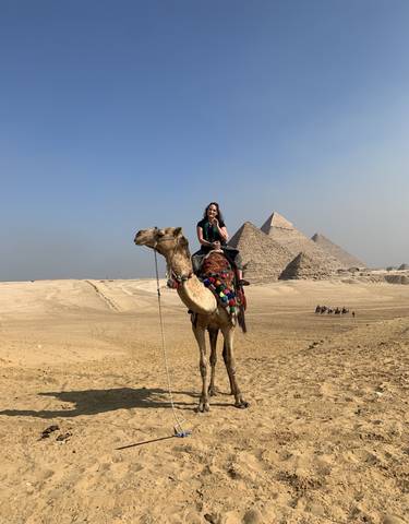 Person riding a camel in front of the pyramids.