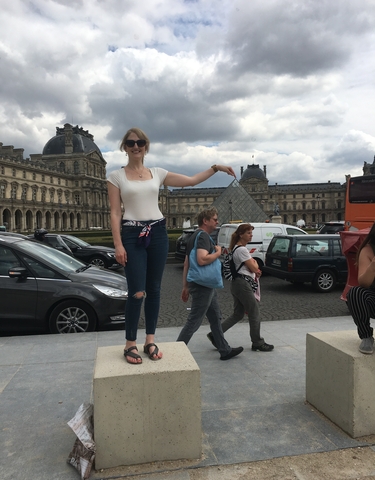 Woman posing as if holding the tip of a glass pyramid with people around.