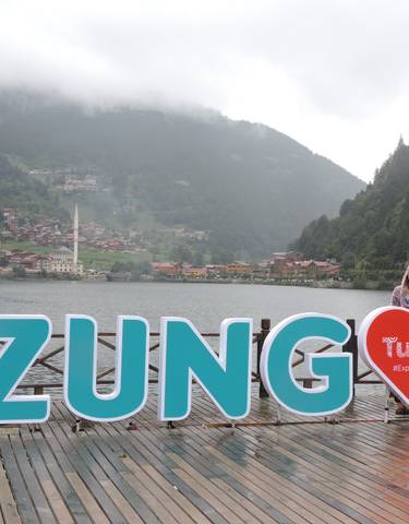 A person posing next to a large Uzungöl sign with a village and nature backdrop.