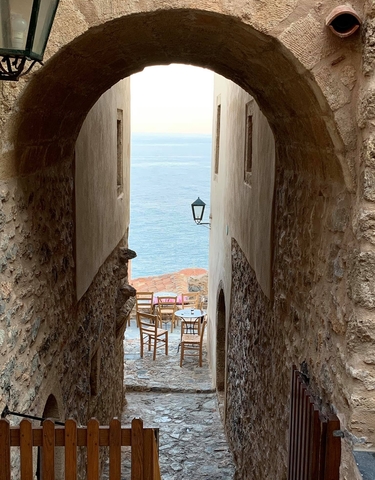 A narrow passageway between buildings leading to a view of a calm ocean with a table and chairs set.