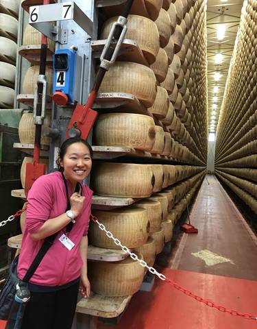 Woman posing next to large cheese wheels in a storage room.