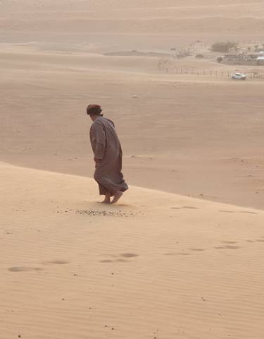 A person walking barefoot across sand dunes with scattered settlements.