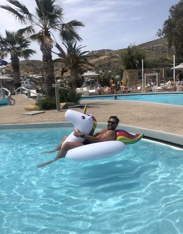 Man enjoying a pool ride on a unicorn float with a resort in the background.