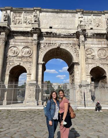 Two people standing in front of a large ancient arch with detailed carvings.