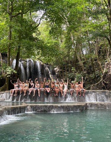 Group of people sitting in front of a waterfall, raising their hands.