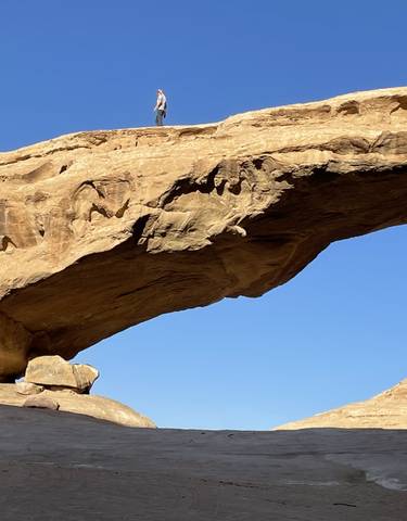 Person standing on top of a large natural rock arch in a desert landscape.