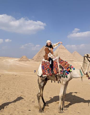 A person riding a camel with pyramids in the background under a clear sky.