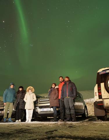 People standing under the northern lights with cars nearby.