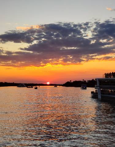 Sunset over a river with boats and silhouettes of people on deck.