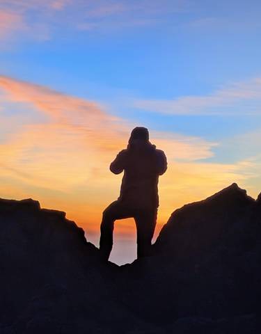 Silhouette of a person photographing the sunset from a mountain peak.