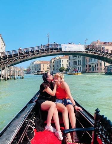 Two women on a gondola in Venice with a bridge and buildings in the background.