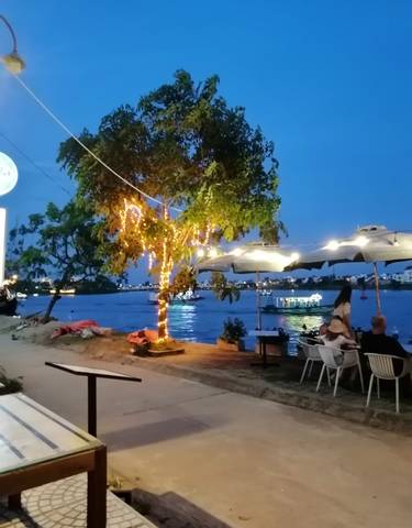 Outdoor dining by a river with lit up trees at night.