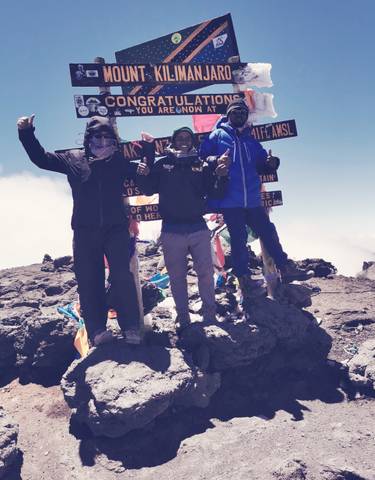 Three people at a mountain summit with flags and signs.
