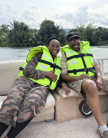 Two people wearing life vests sitting on a boat with greenery in the background.