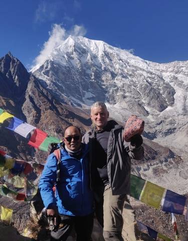 Two people posing in the mountains with prayer flags and snow-covered peaks.