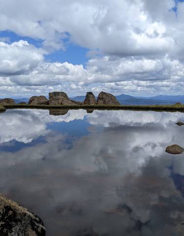 A small body of water reflecting a cloudy sky surrounded by rugged terrain.