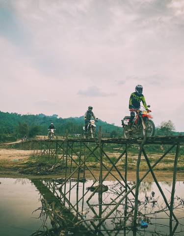 Motorbikes crossing a rustic wooden bridge over a reflective water body.
