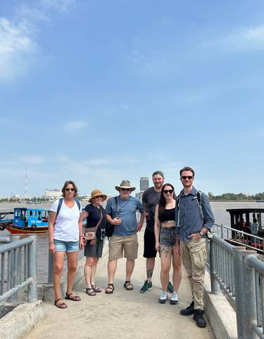Group of people standing on a pier with boats on the river and buildings in the background.
