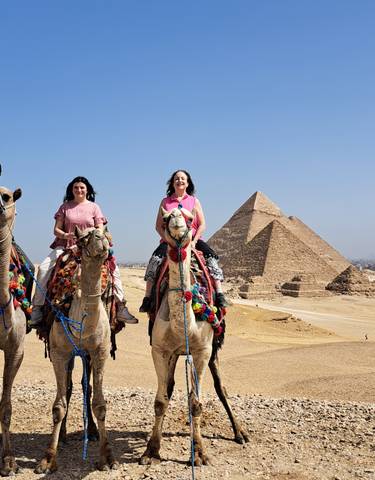 Three people riding camels with the pyramids in the background.