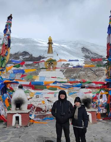 Two people in front of a colorful stupa with prayer flags and snowy mountains in the background.