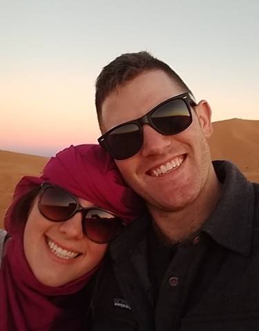 Couple smiling in a desert landscape with sunset in the background.