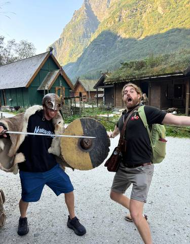 Two men playfully posing with sword and shield in a village setting.