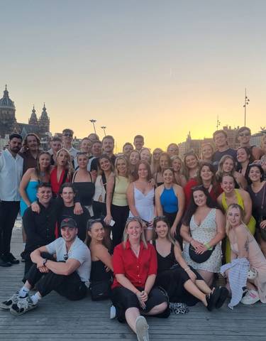 Large group posing by water during sunset with cityscape.