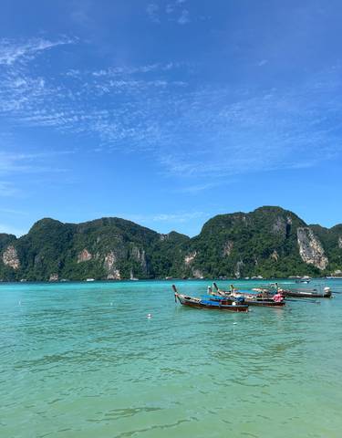 Panoramic view of long-tail boats on turquoise water with green mountains.