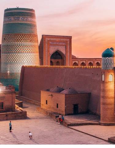 People walking in front of Khiva's iconic minaret and city walls at sunset.