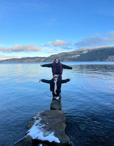 Two people posing by a lake in a scenic landscape.