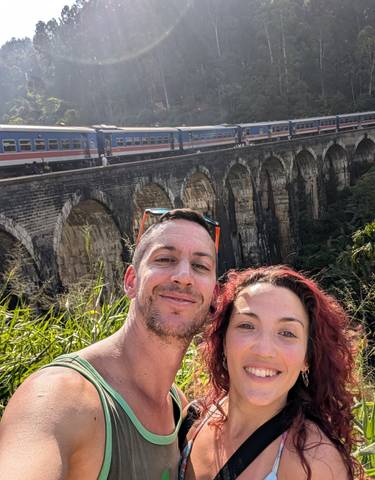 Selfie of a couple with a train on a bridge in the background.